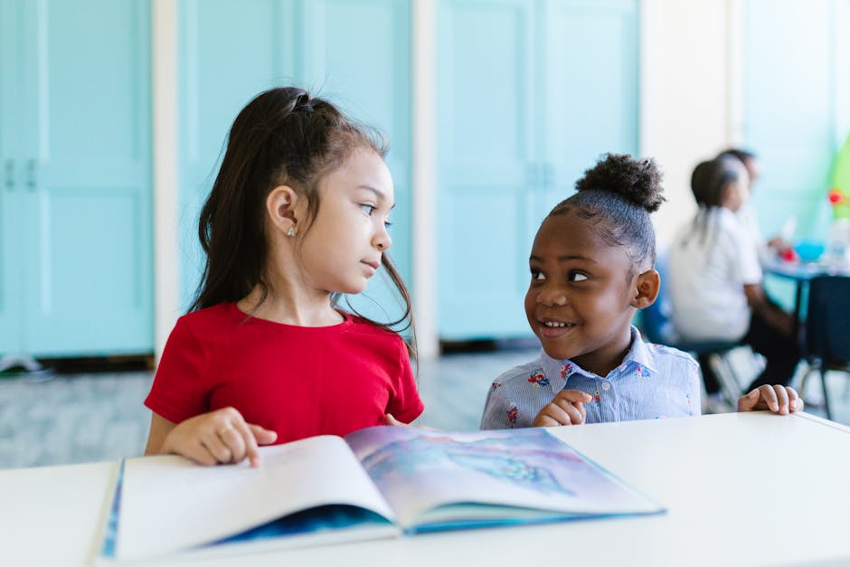 Two young children in a classroom interact while reading a book together