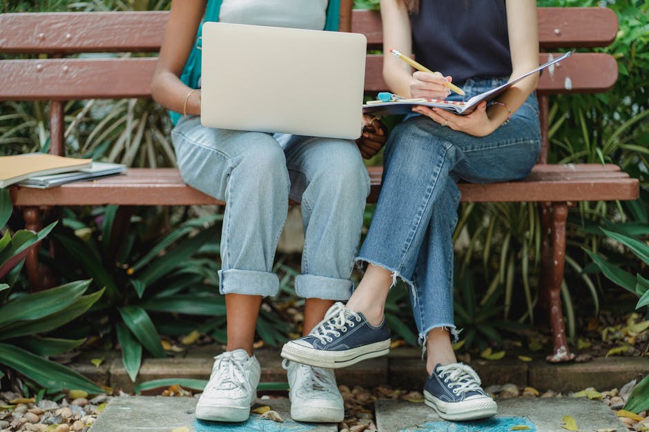Two young women collaborating on a park bench with a laptop and notebook during summer