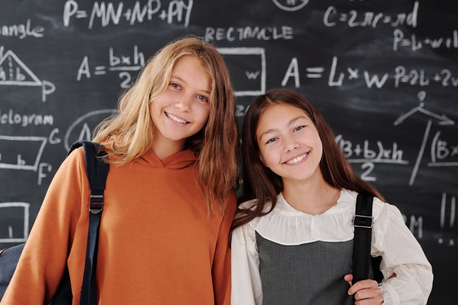 Two cheerful girls stand in a classroom with a math-covered chalkboard, ready for learning